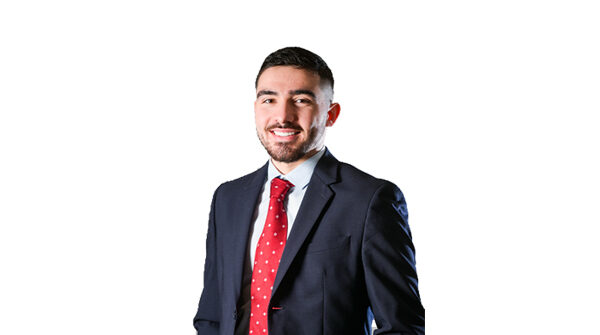 Portrait d’un jeune homme, alumni de l'ESTA Belfort, souriant, vêtu d’un costume bleu marine, chemise claire et cravate rouge à motifs, posé devant un fond gris neutre.