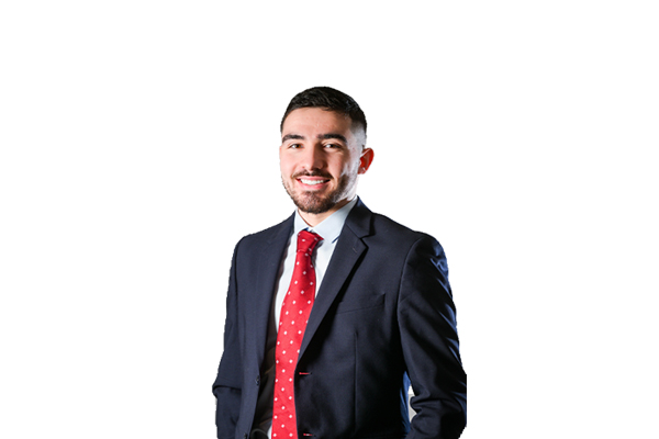 Portrait d’un jeune homme, alumni de l'ESTA Belfort, souriant, vêtu d’un costume bleu marine, chemise claire et cravate rouge à motifs, posé devant un fond gris neutre.