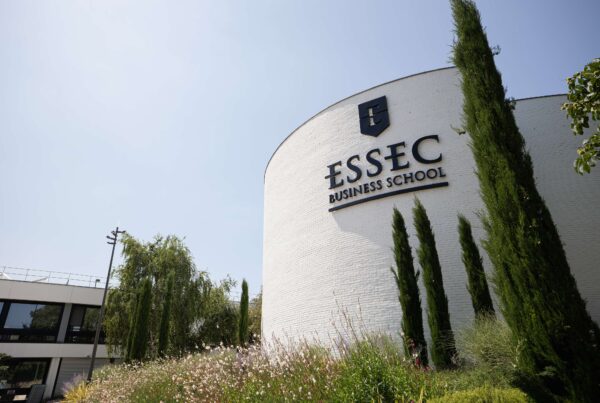 Façade blanche arrondie du bâtiment de l'ESSEC Business School à Cergy, ornée du logo noir de l'école, entourée de verdure et de cyprès sous un ciel bleu.