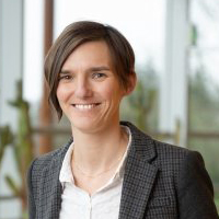 Une femme en tenue professionnelle, souriante, se tient debout dans un bâtiment lumineux avec de grandes baies vitrées.